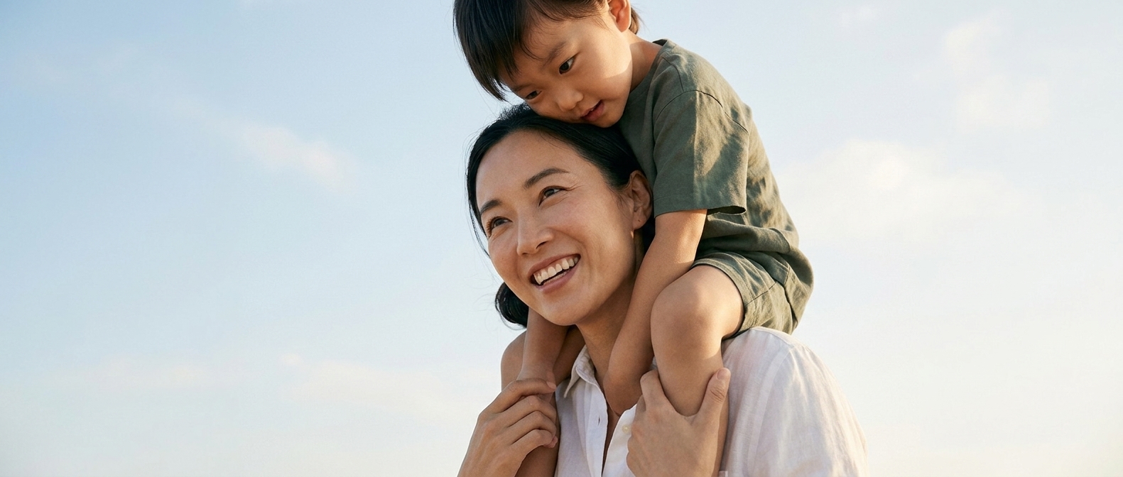 A mother carrying her young son on her shoulders against a bright sky
